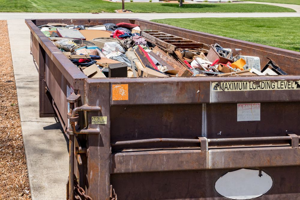 Full, rusty dumpster overflowing with debris on concrete.
