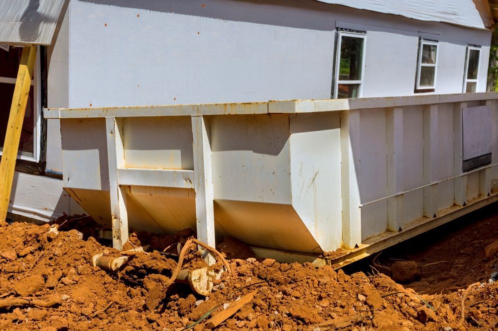 White dumpster next to a house under construction; sitting on reddish-brown dirt.
