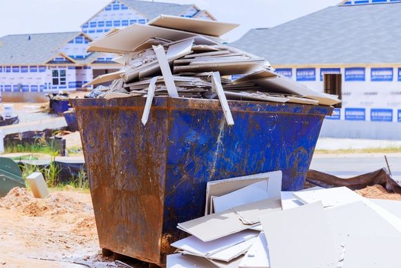 Blue construction dumpster overflowing with drywall, in front of houses under construction.