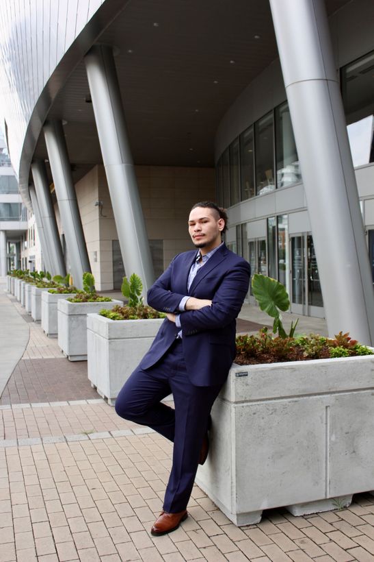 A man in navy suit leaning against structure with arms crossed