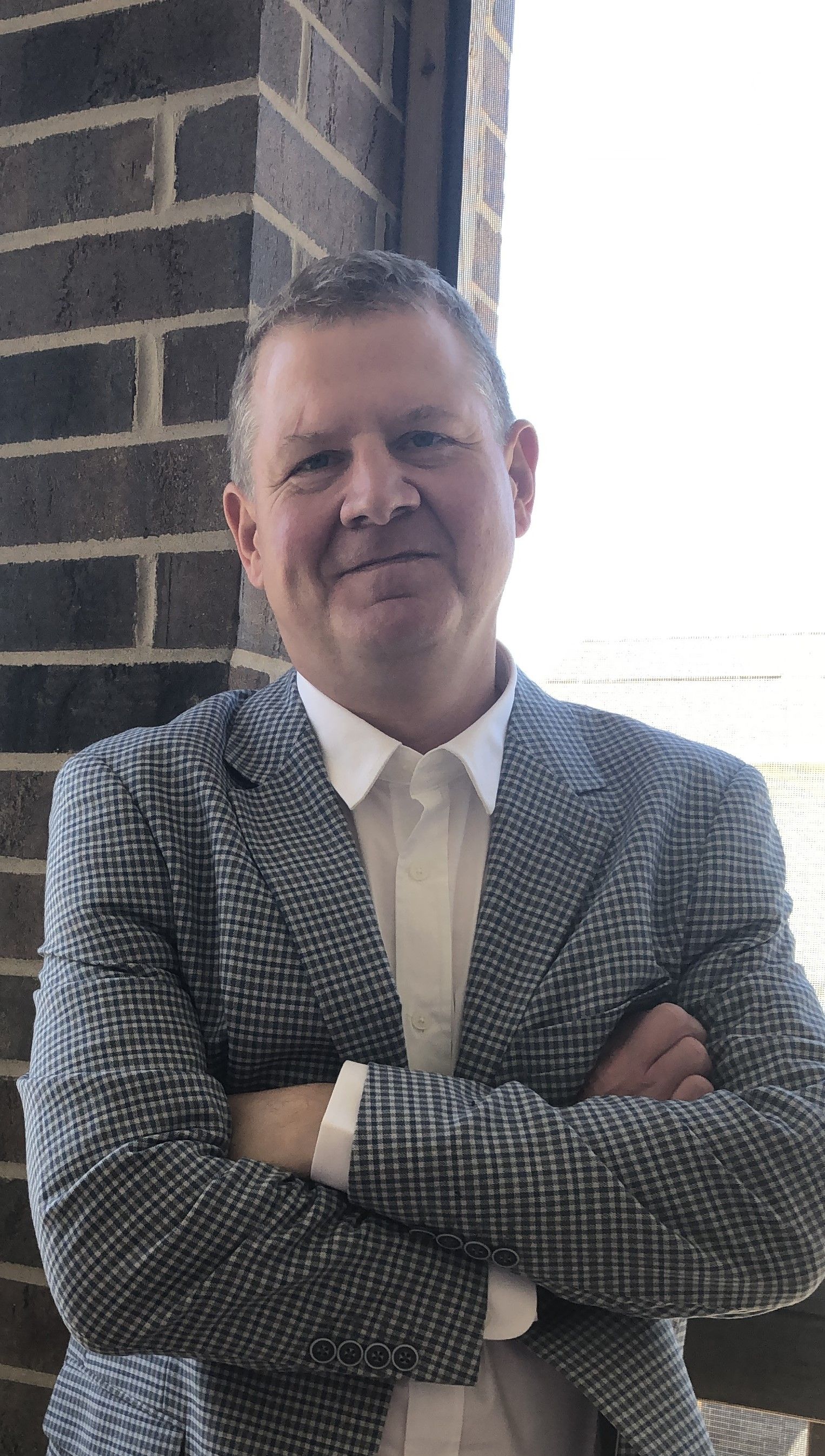 A man in a suit is standing with his arms crossed in front of a brick wall.