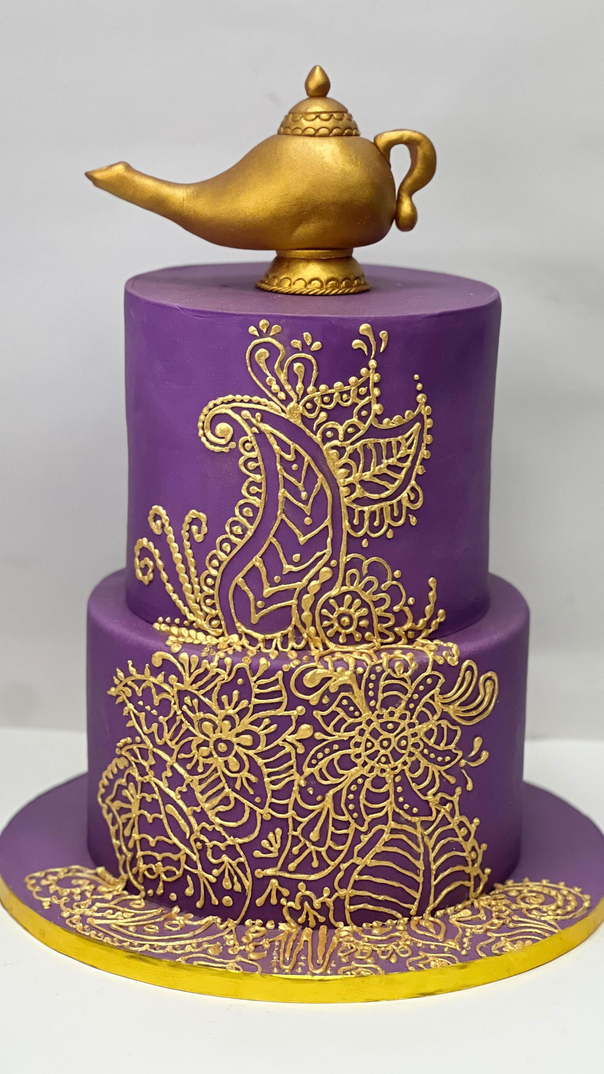 Torta viola a due piani con motivo paisley dorato e lampada genio dorata sulla parte superiore.