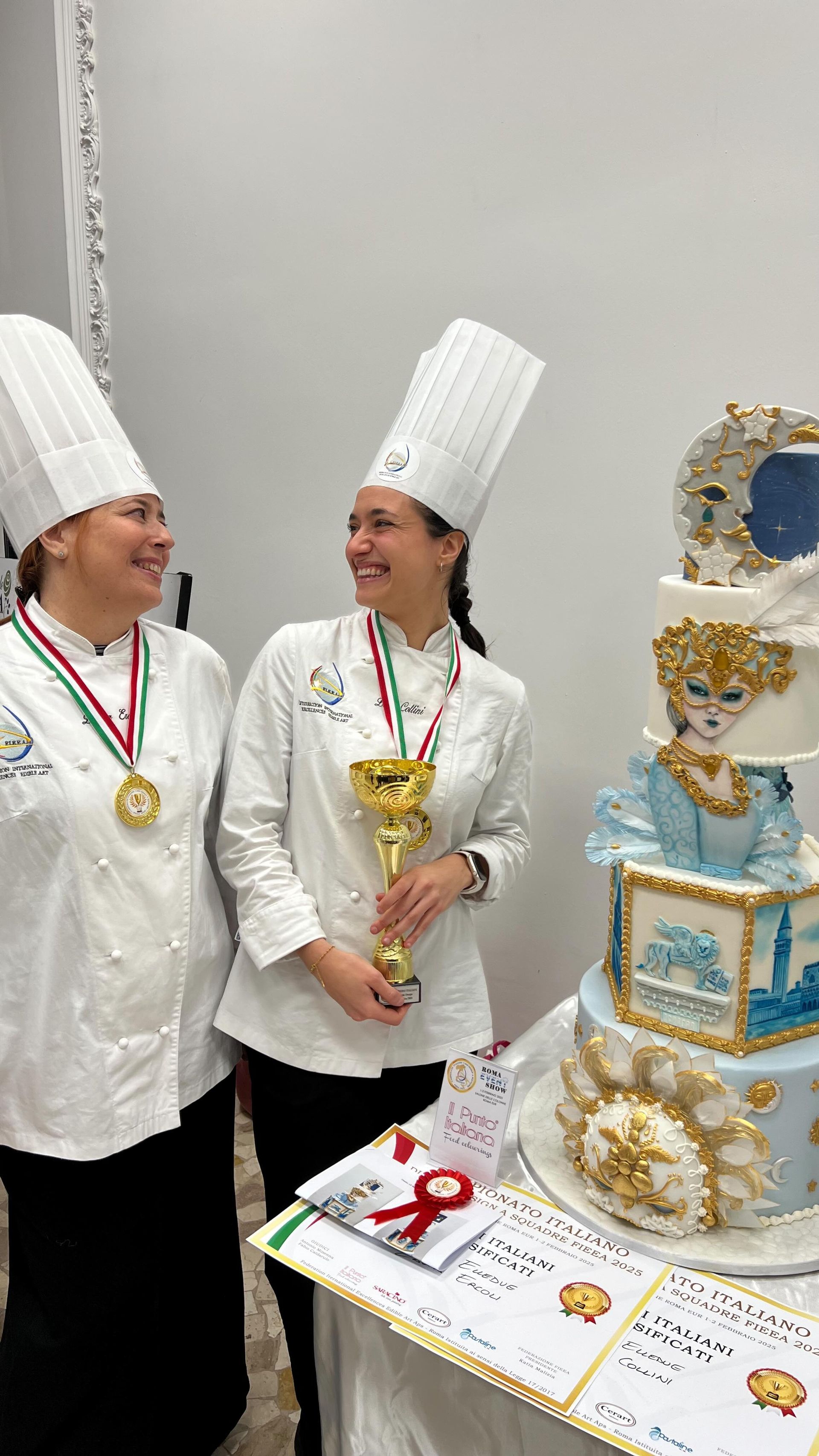 Due chef in uniforme bianca sorridono, uno dei quali tiene in mano un trofeo, accanto a una torta decorata.