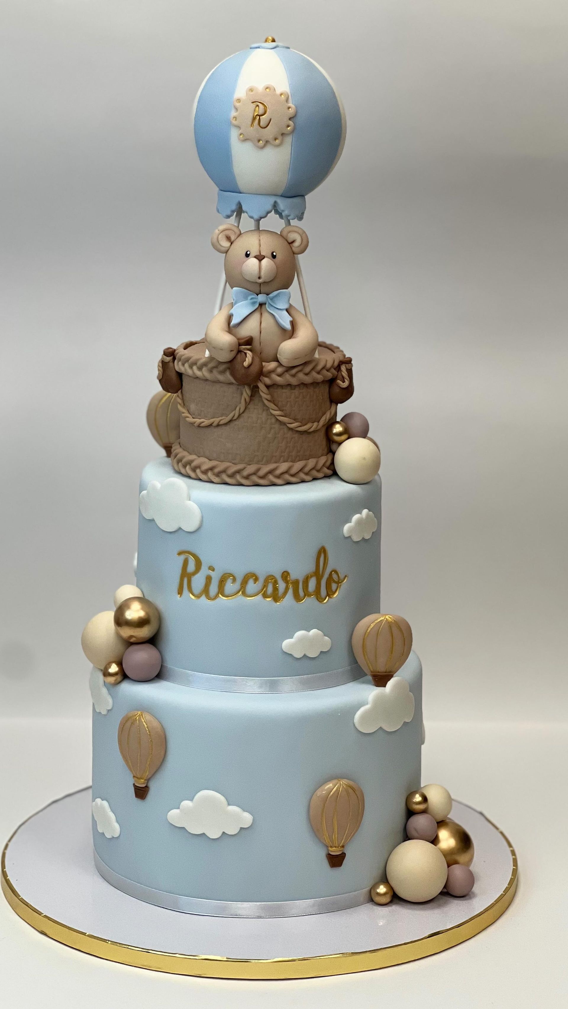 Torta per baby shower a due piani con orsetto in mongolfiera, dettagli azzurri e dorati e il nome