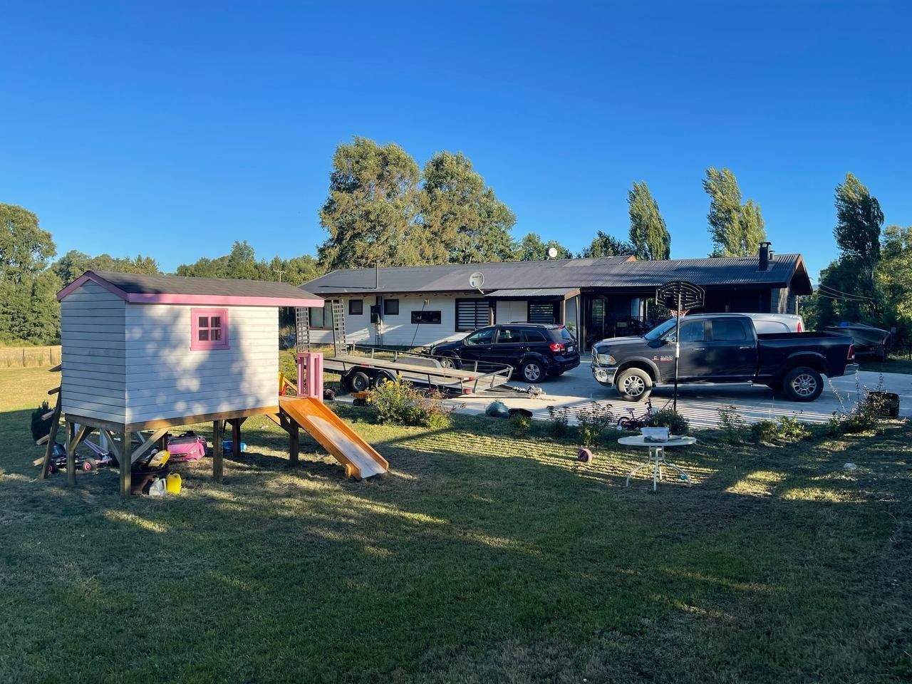 Una casa con sala de juegos, vehículos y un remolque sobre un césped verde bajo un cielo azul claro.