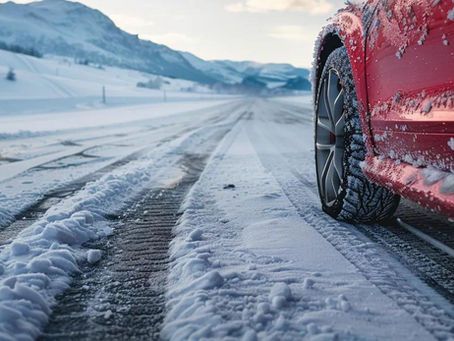 Red car driving on a snowy road with tire tracks, mountains in background.