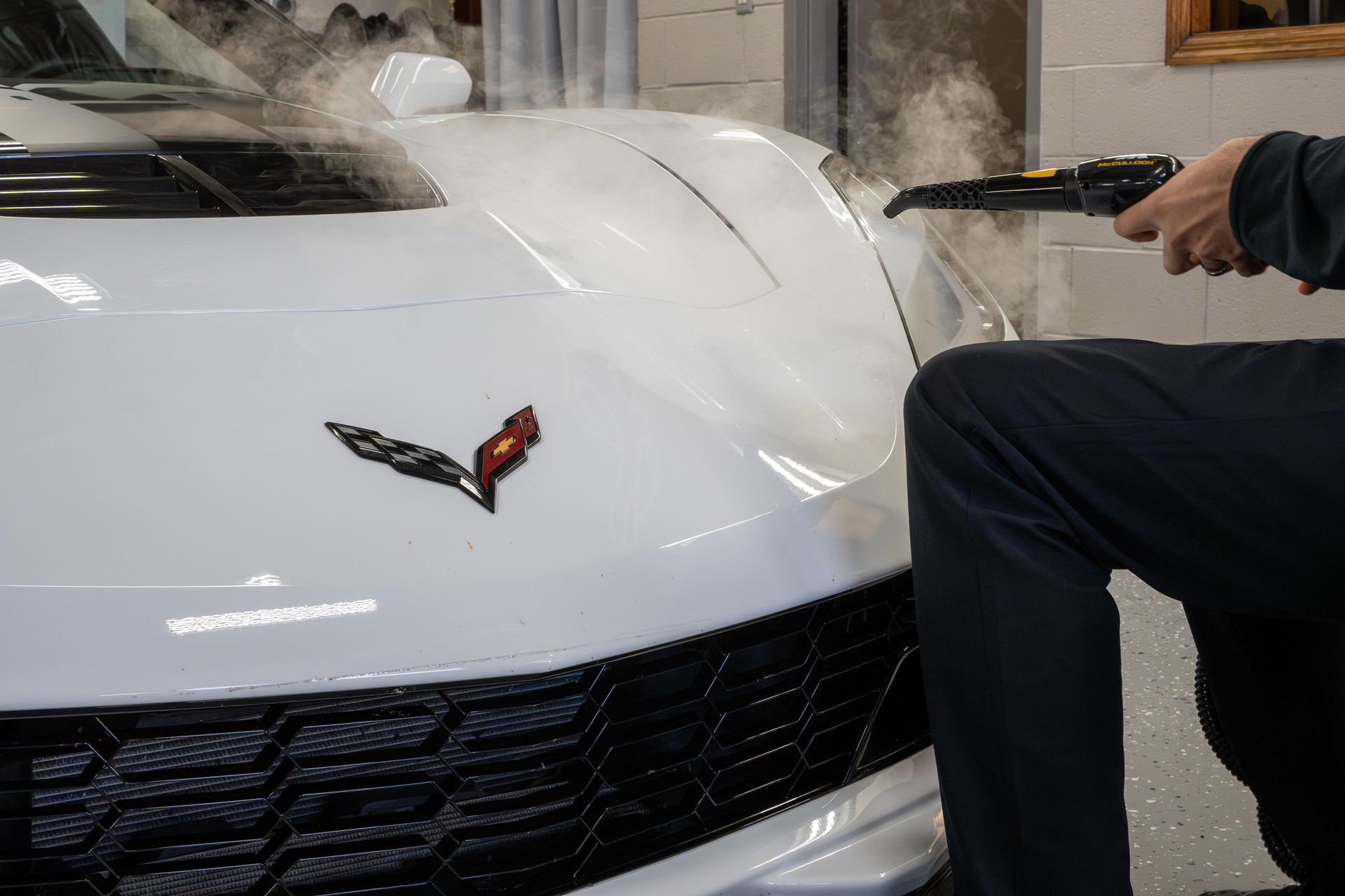 Person steam cleaning a white Corvette's hood, black grill and logo visible.