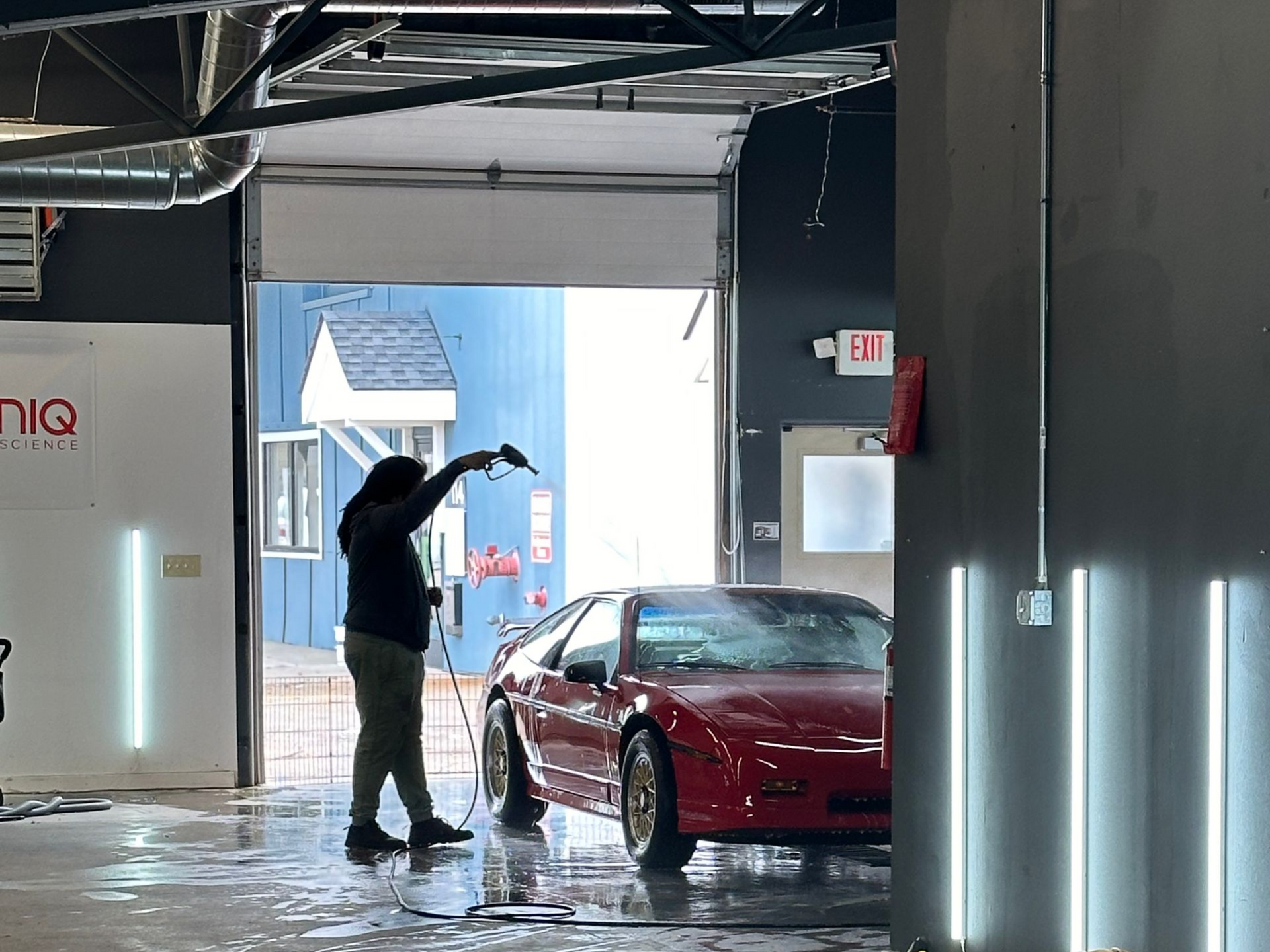 Person washing a red sports car inside a garage; blue building in the background.