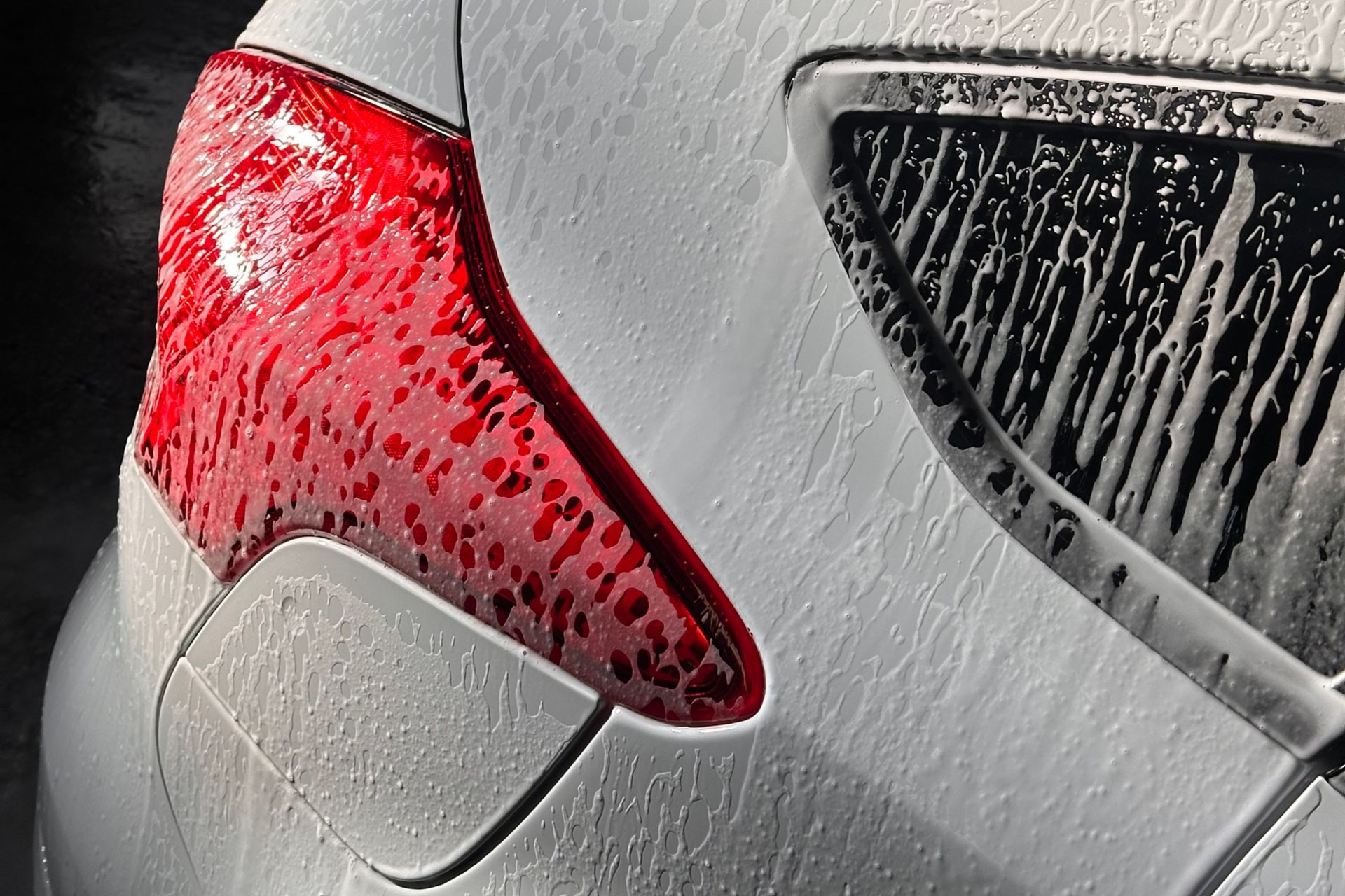 A white car covered in foamy soap at a car wash. Headlight and grill are visible.