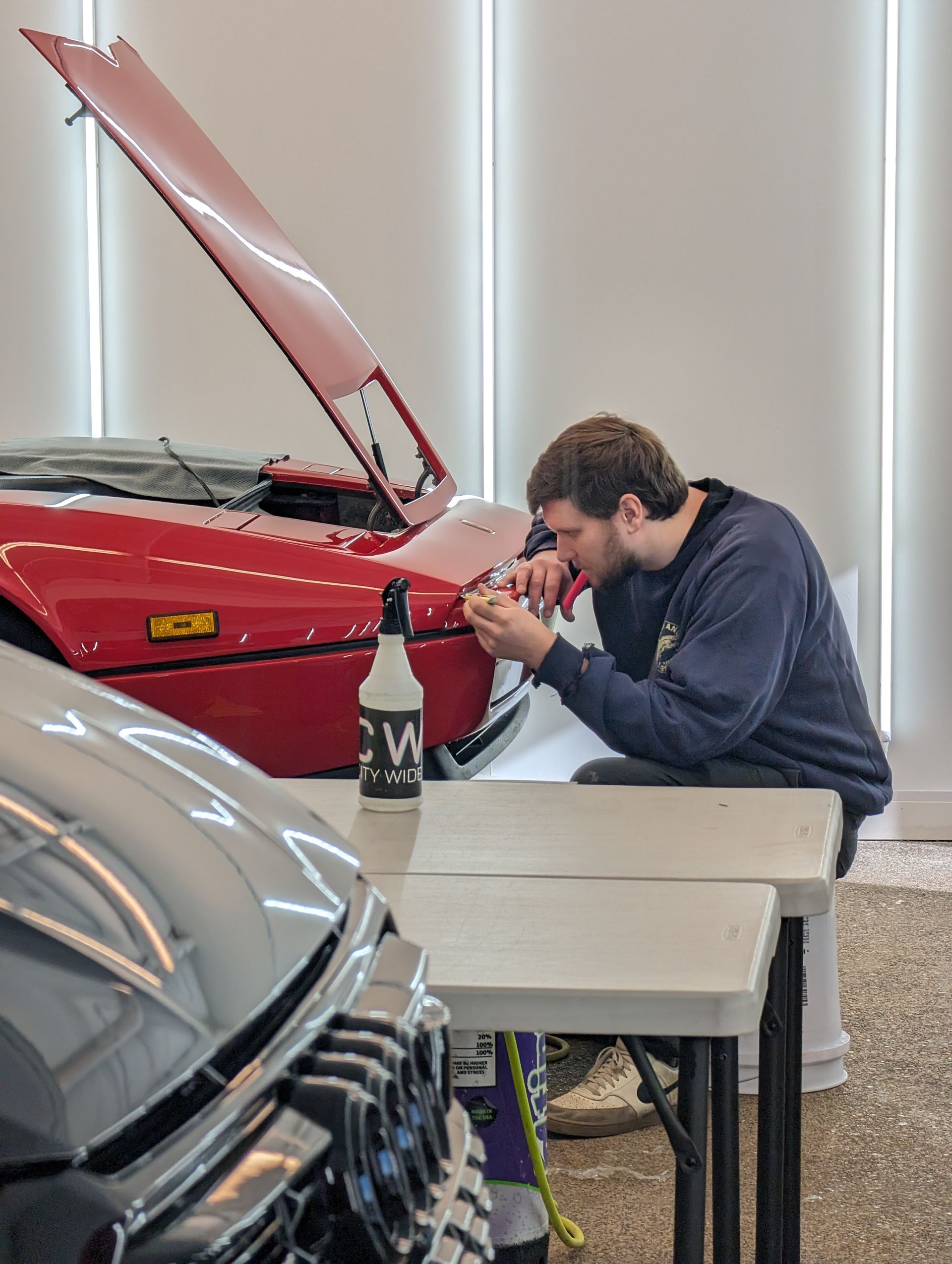 Man working on a red car with hood up, seated at a table. Interior shot.