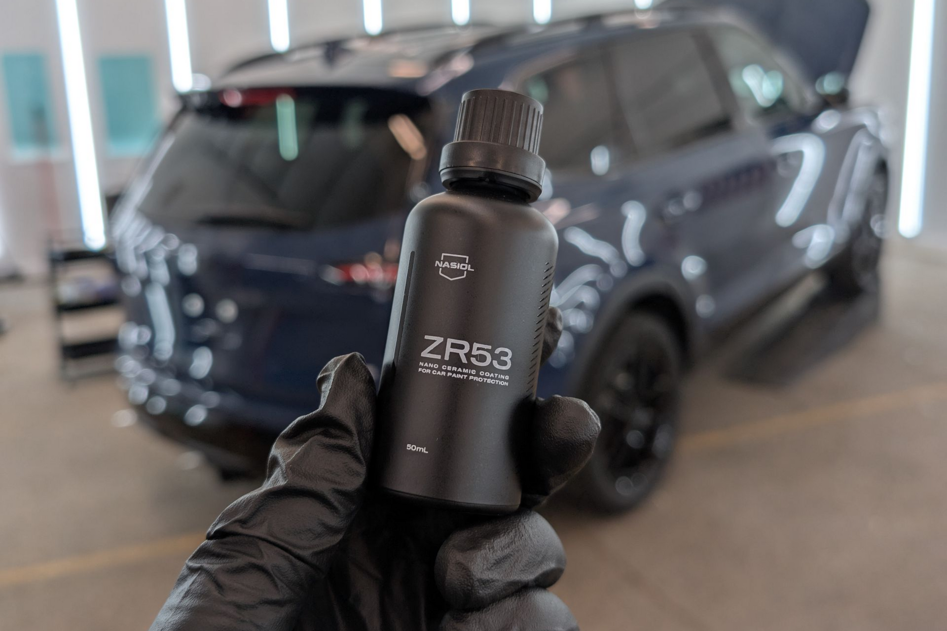 Person in black gloves applying coating to a black car in a workshop.