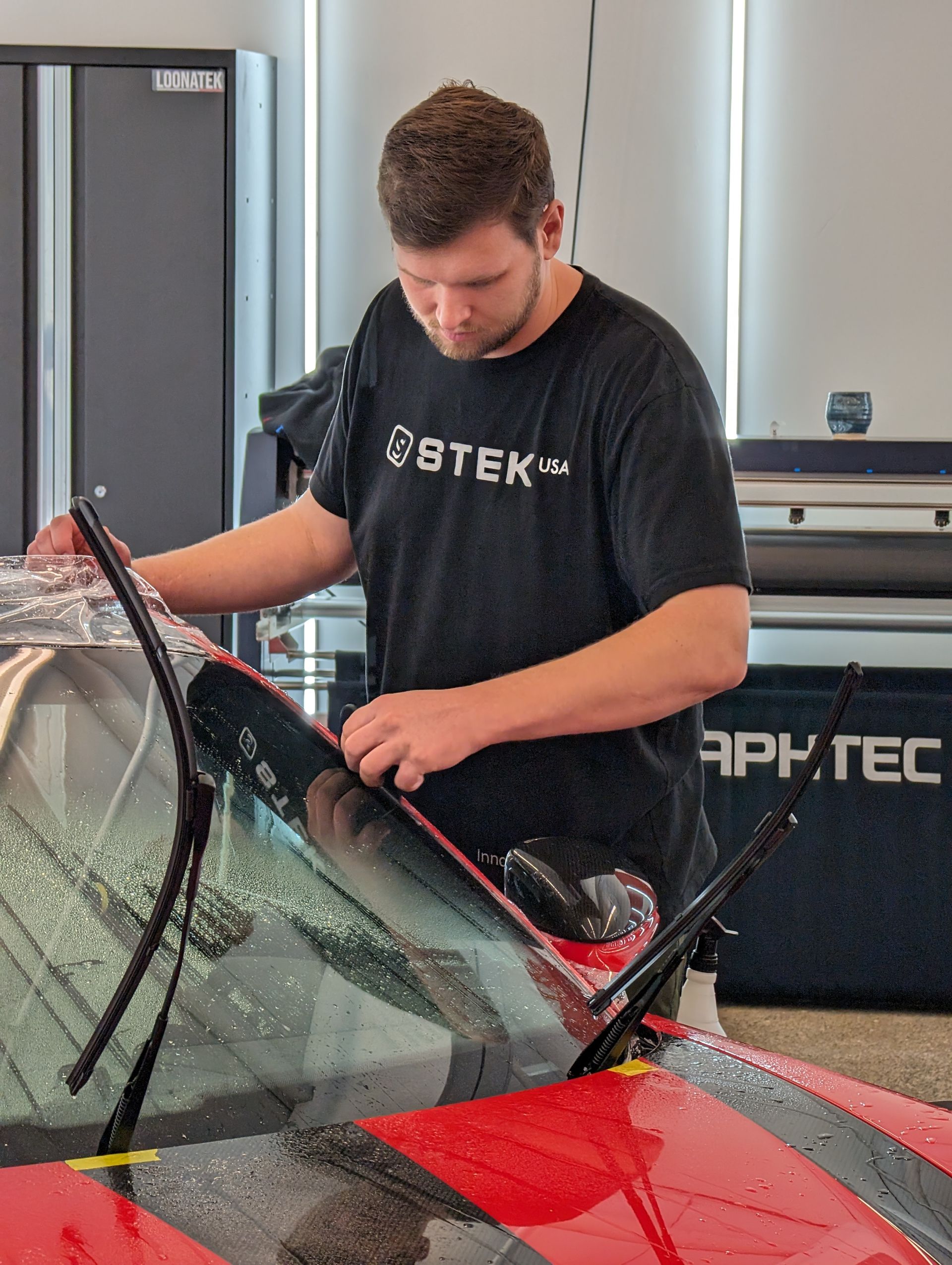 Man in black shirt applying film to a red car windshield inside a shop.