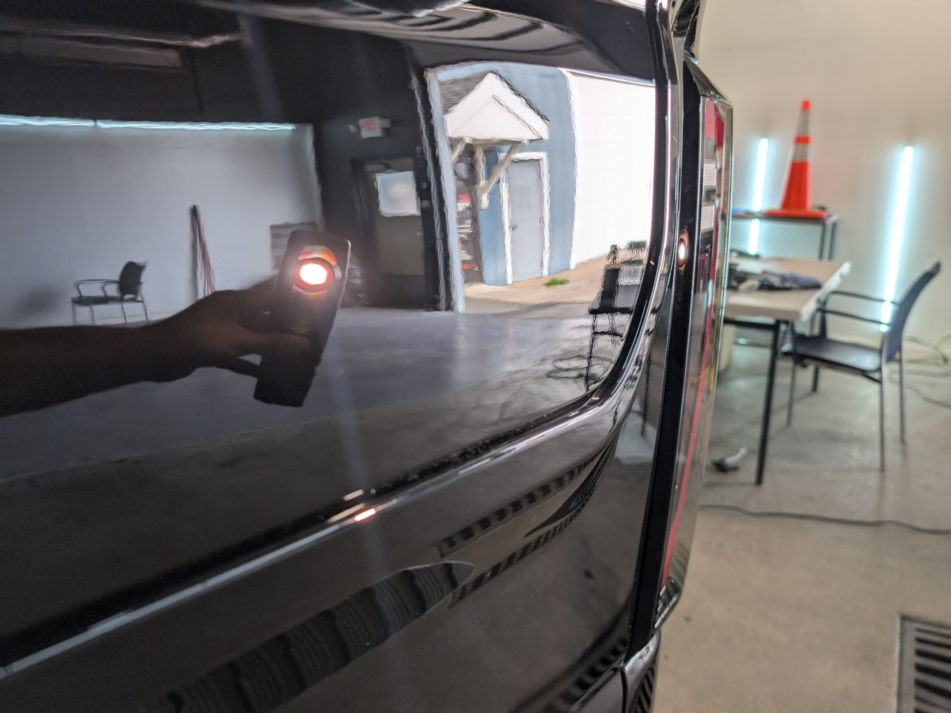 Black car's polished surface reflects a shop interior with a person holding a light, checking the finish.