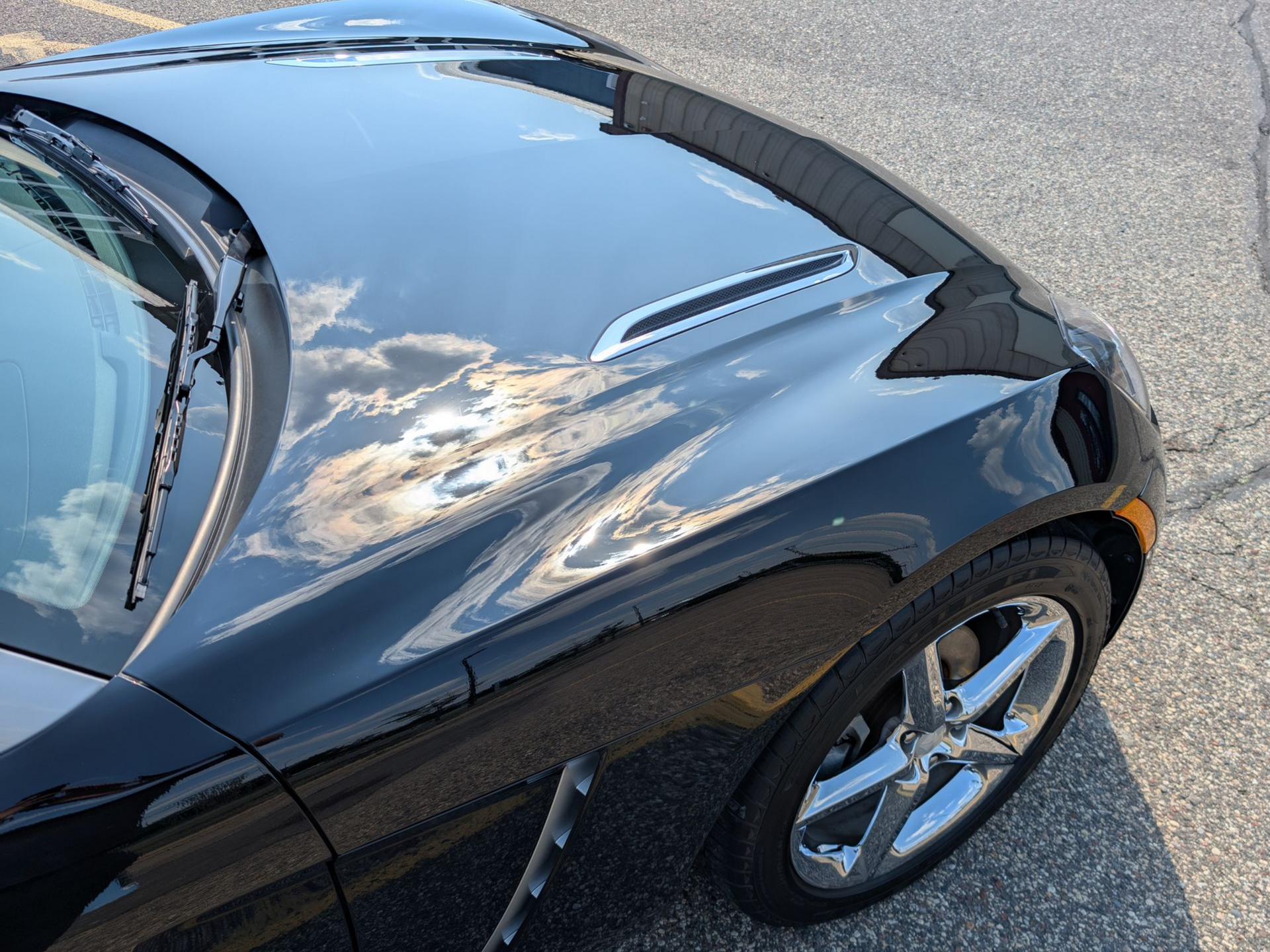 Black sports car hood, reflecting clouds and sunlight, parked on asphalt.