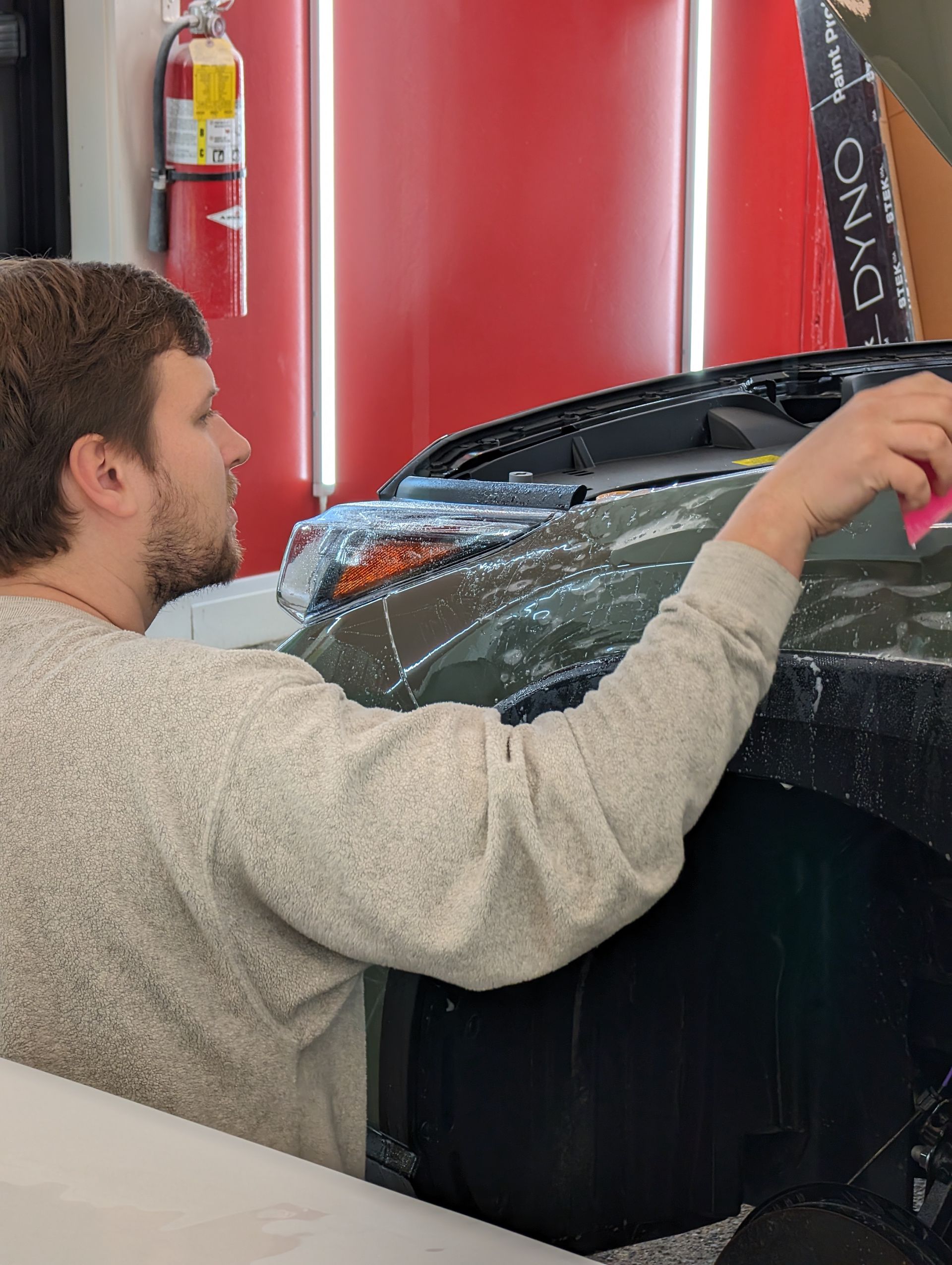 Man applying film to a car's hood in a shop. Red wall, fluorescent lights, and fire extinguisher are visible.