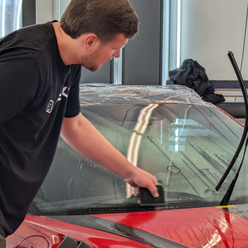A close-up of a car's windshield, reflecting overhead lights. Wipers are visible; a green sticker is also on the glass.