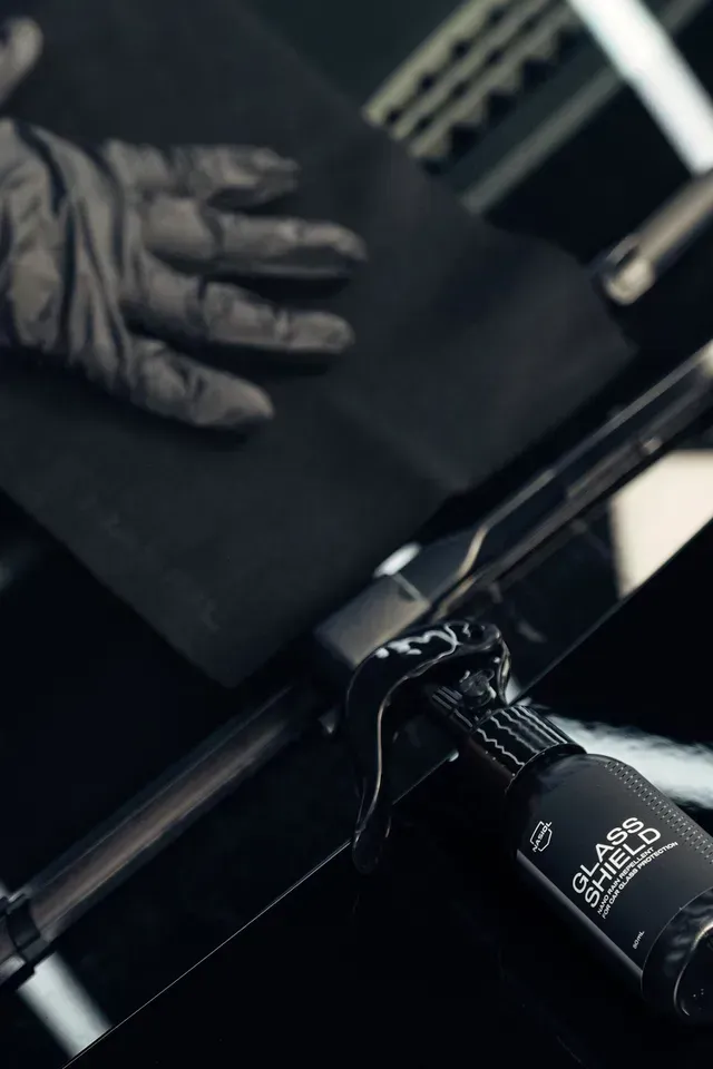 A gloved hand wipes a dark surface next to a bottle of Glass Shield professional coating product.