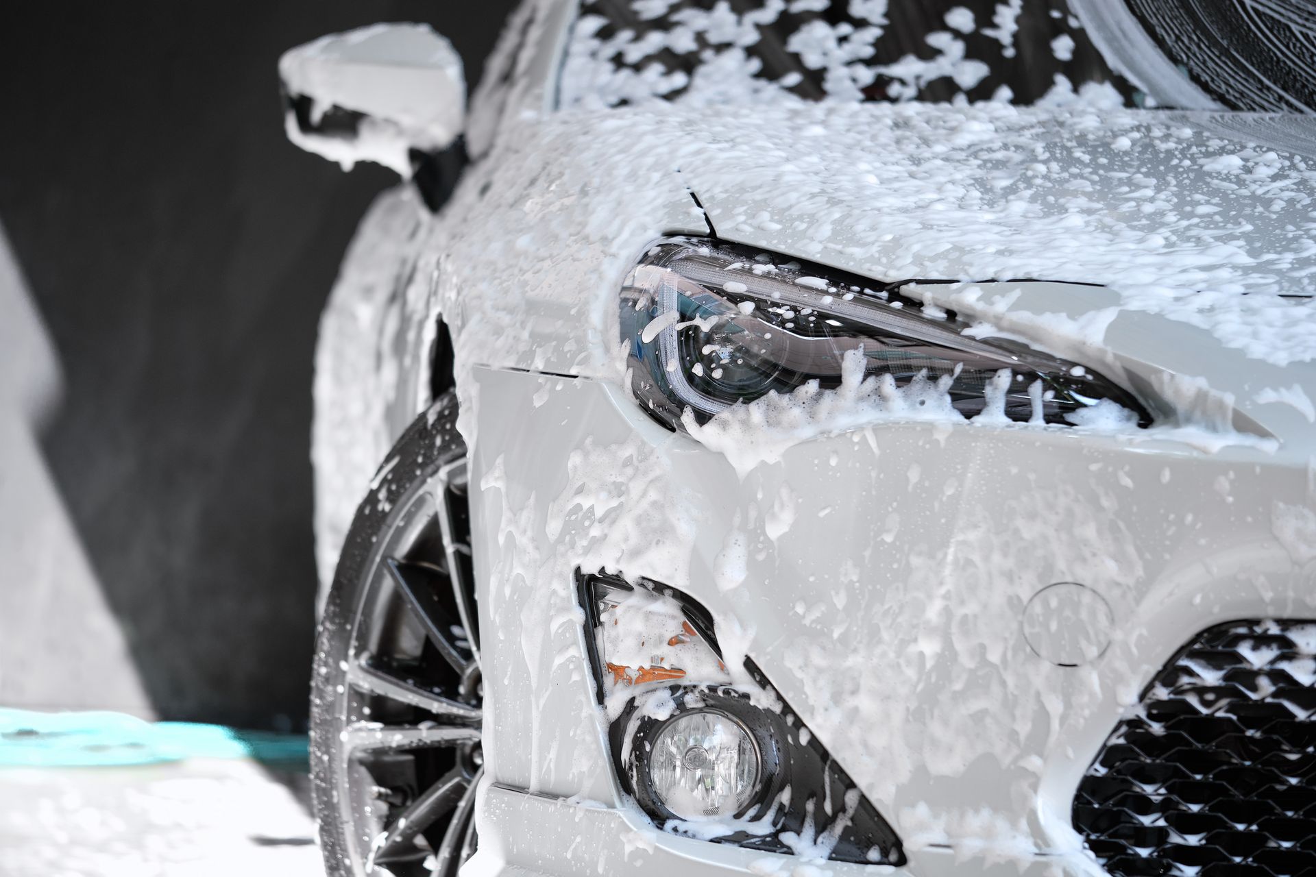 A white car covered in foamy soap at a car wash. Headlight and grill are visible.