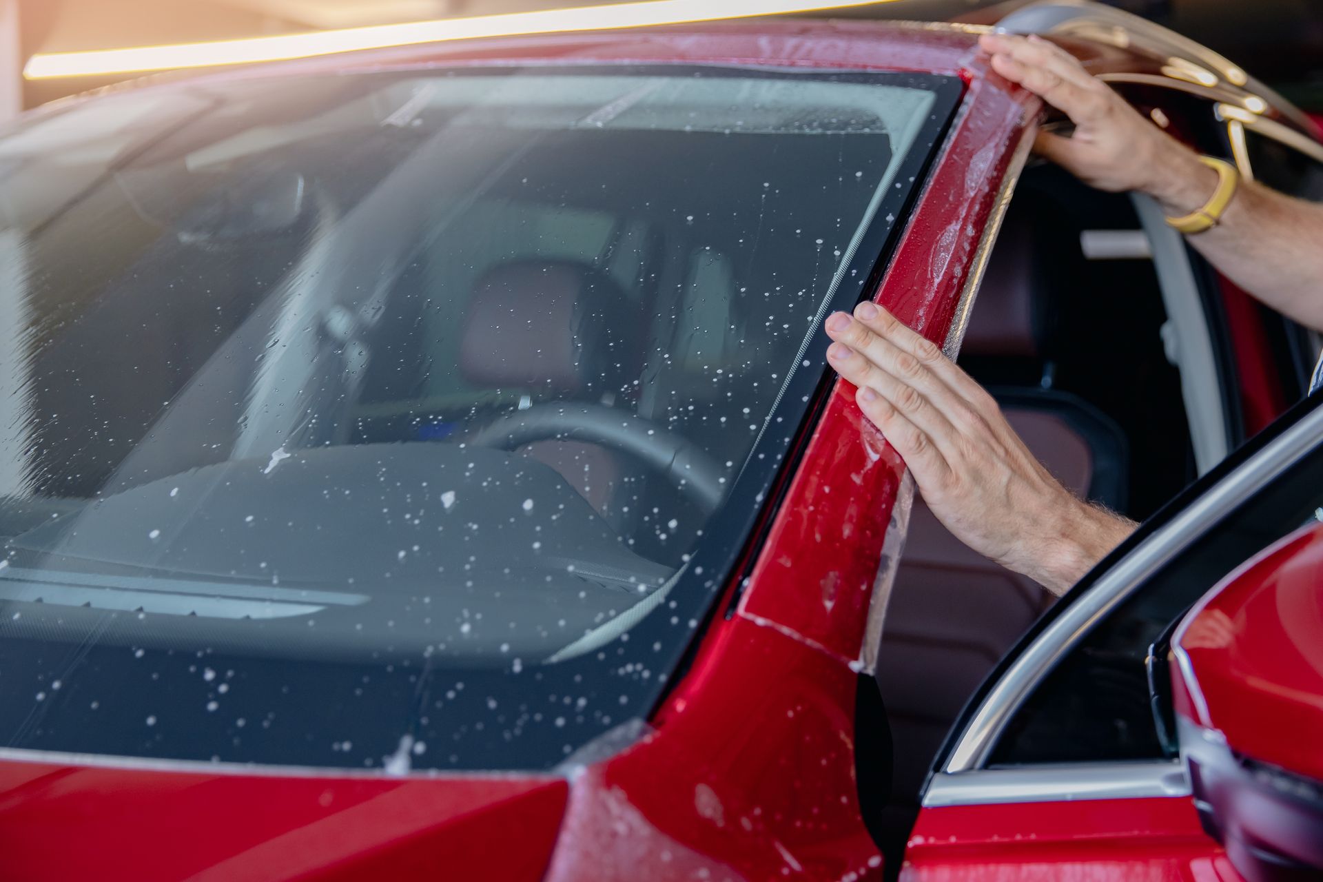 Hands cleaning a red car's windshield and frame with soapy water at a car wash.