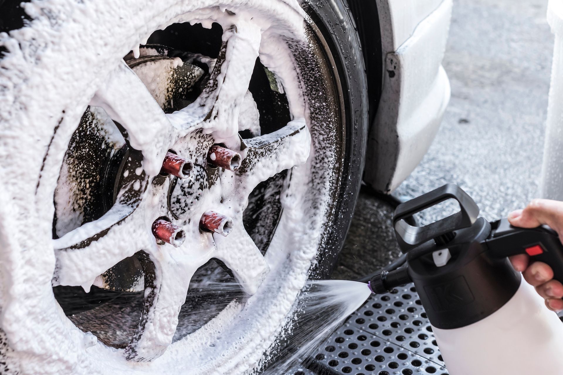 Foamy car wheel being sprayed with cleaning solution by a person, on a gray surface.