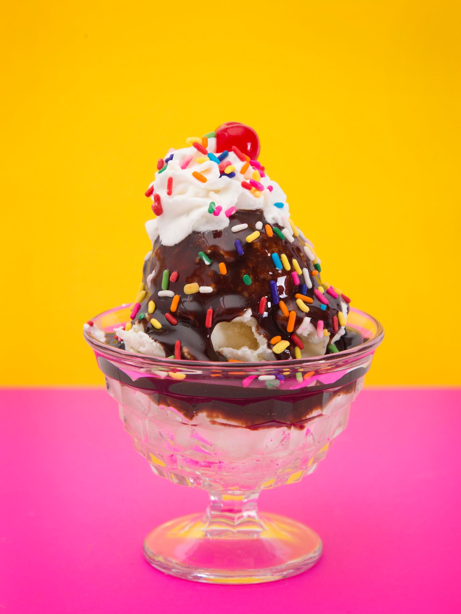 A sundae with chocolate sauce , whipped cream , and sprinkles on a pink and yellow background.