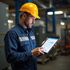 Man in yellow hard hat and blue work shirt using a tablet in a factory setting.