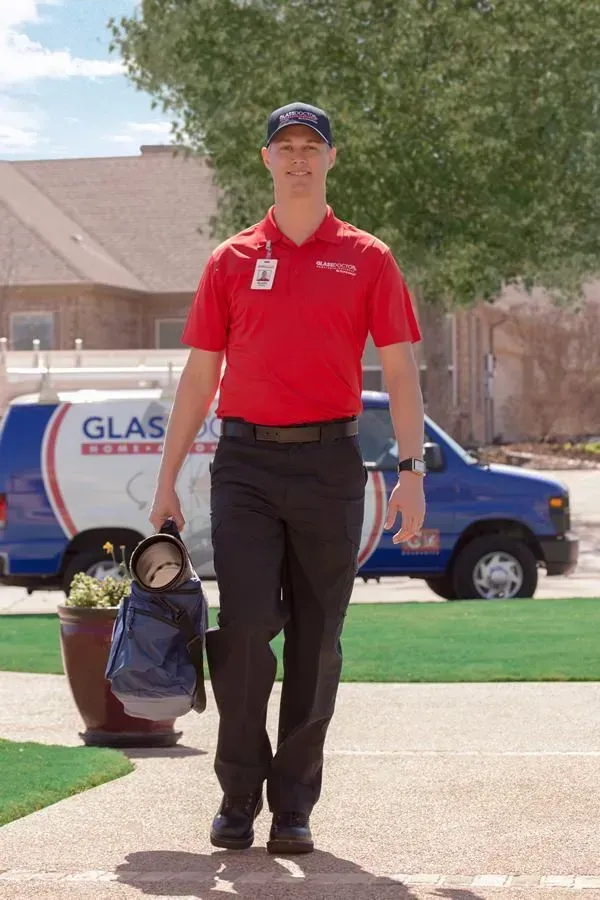A person in a red shirt and black pants walks toward the camera, holding a tool bag. A van is in the background.