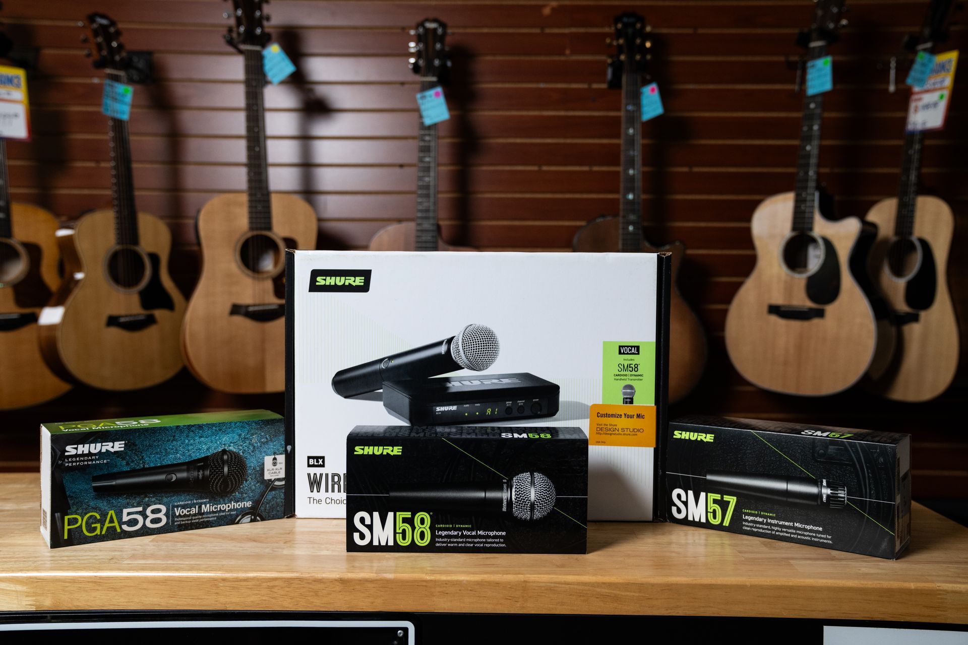 Microphones and guitar boxes on a wooden counter with guitars on a wood-paneled wall.