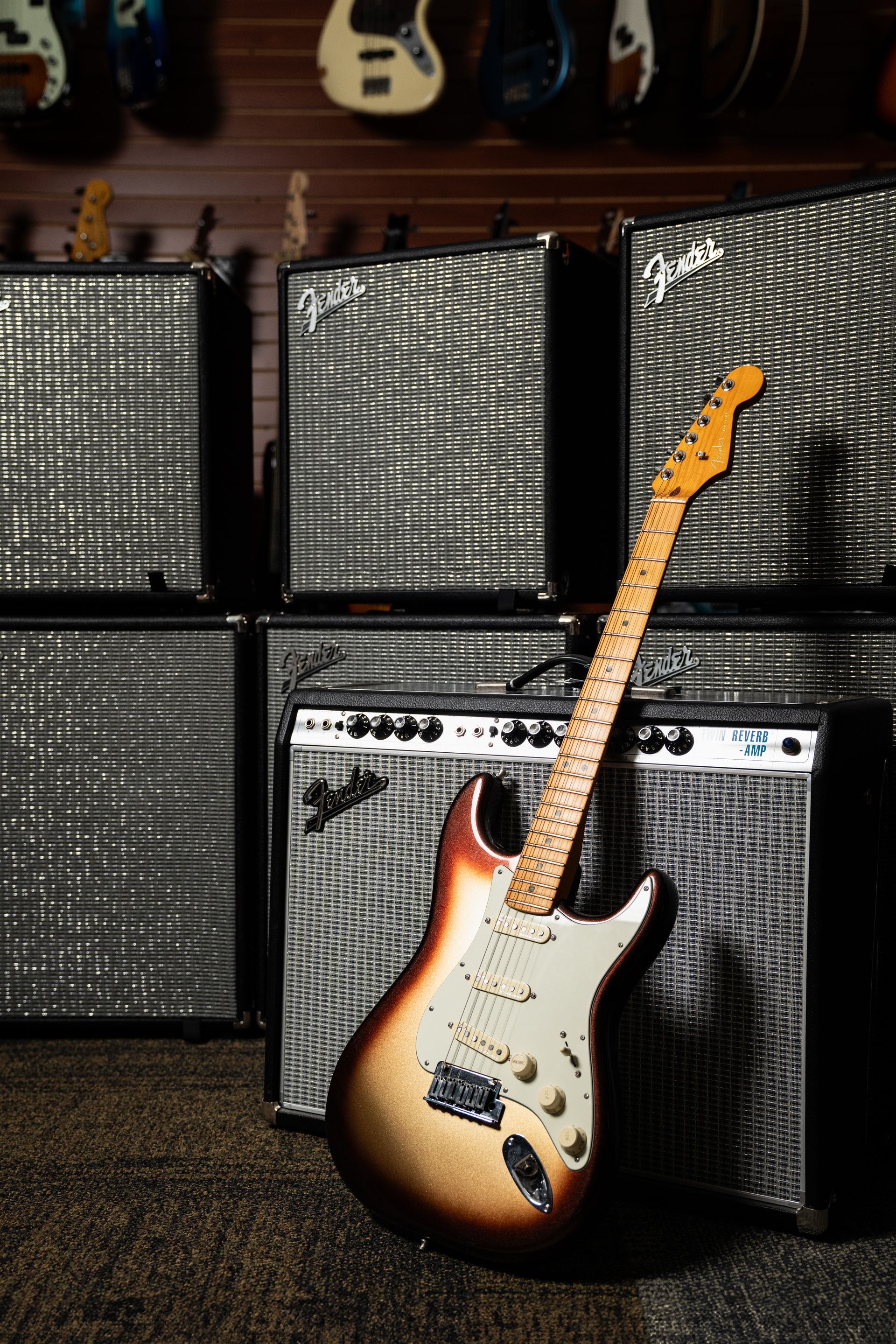 Guitar leans against amplifiers in a music store. Sunburst Stratocaster, silver amp grills, wood-paneled wall.
