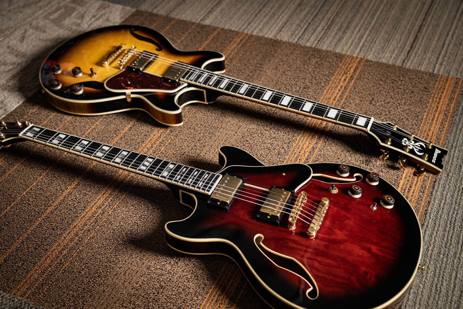 Two semi-hollow body electric guitars, sunburst and red, lying on a textured surface.