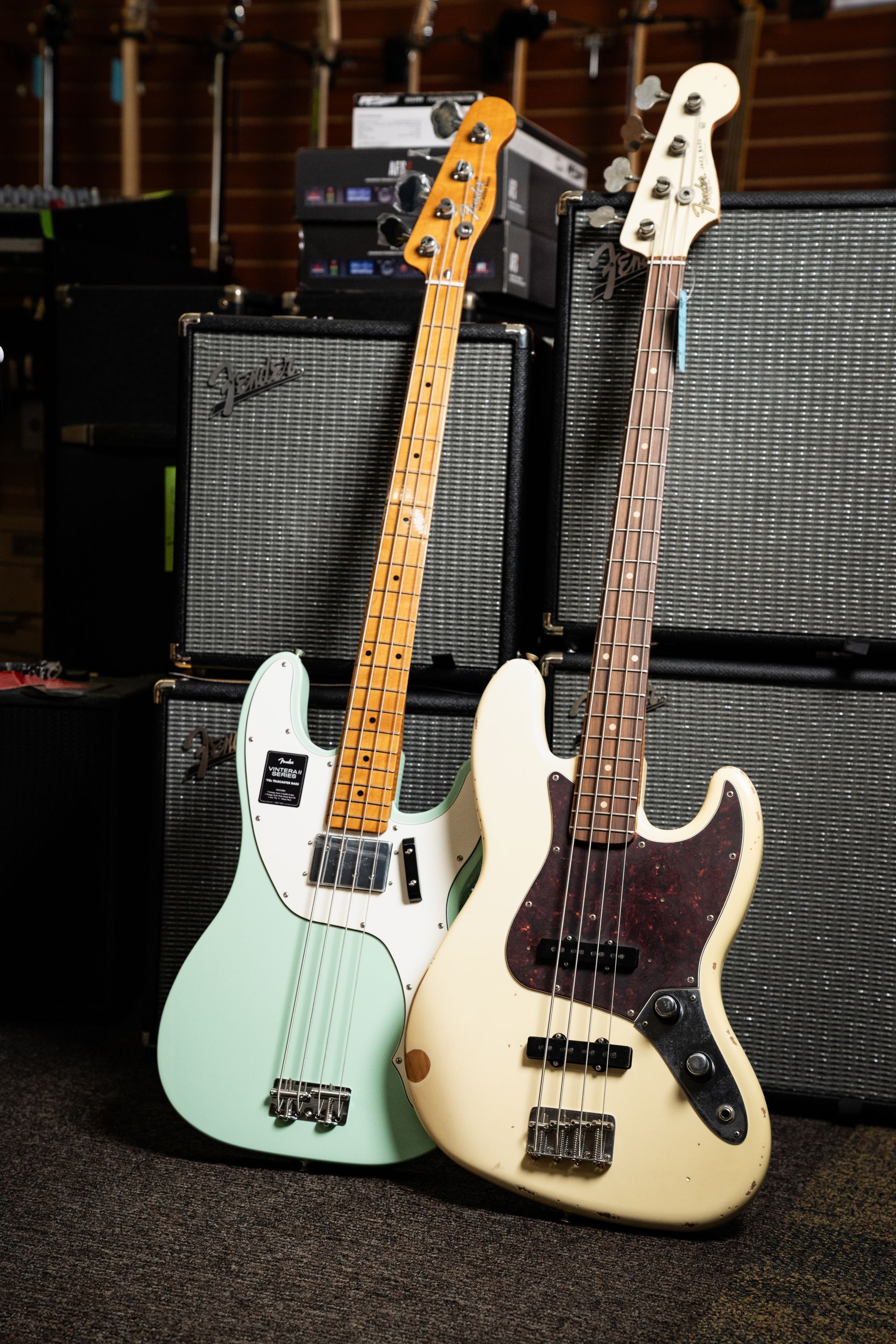 Two Fender electric bass guitars lean against amplifiers in a music store. One is mint green, the other cream.