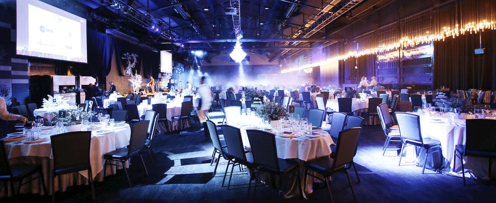 Large event space with round tables set for a banquet. Blue and gold lighting illuminates the room.