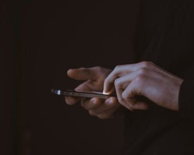 Hands holding a smartphone, fingers tapping the screen; dark setting.