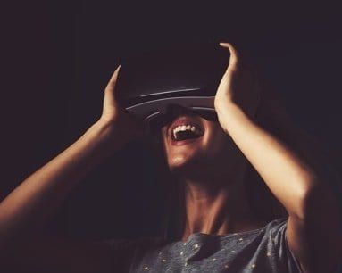 Person wearing VR headset, laughing with hands raised in excitement, dark background.