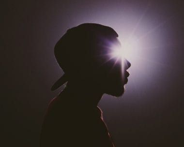 Silhouette of a person wearing a cap, illuminated by bright light shining behind their head.