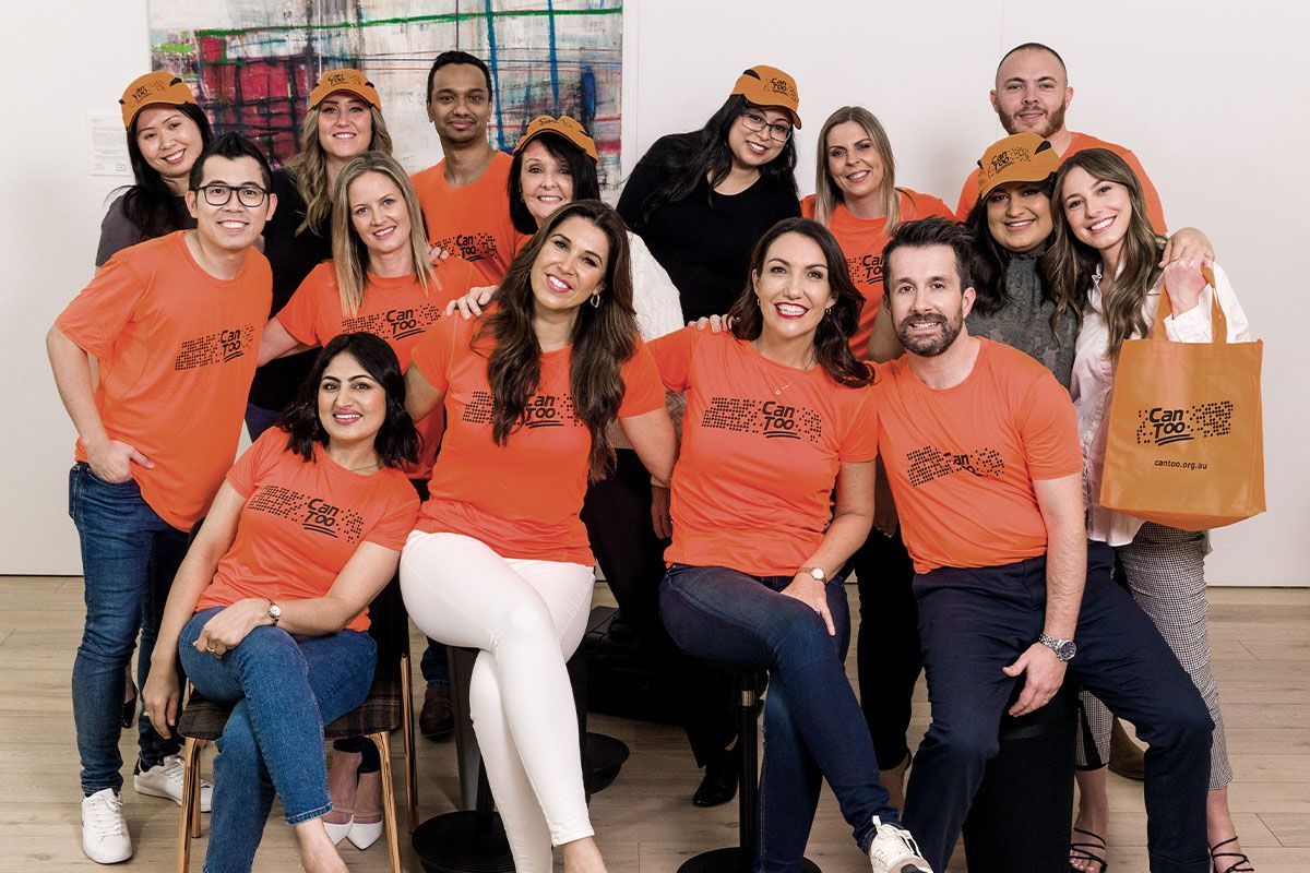 Group of people in orange shirts and hats pose, smiles, some holding a tote bag, indoors.