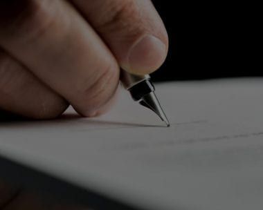 Hand holding a pen, writing on a white paper, close-up shot against a black background.