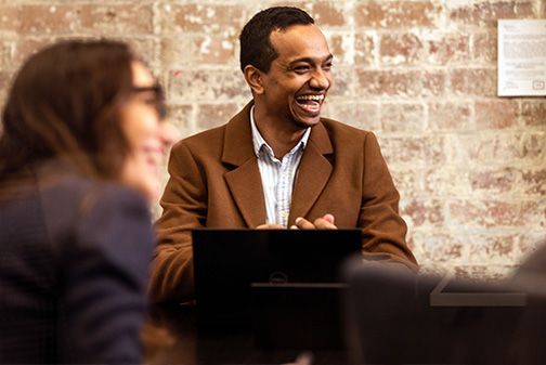 Man laughing, wearing brown coat, at table with laptop, brick wall background.