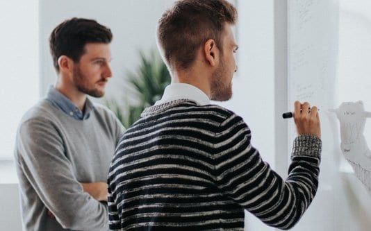 Two men brainstorm at a whiteboard, one writing with a marker, other watching with arms crossed.