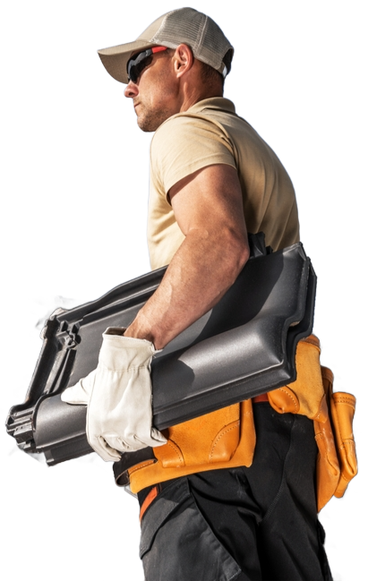 Roofer carrying dark gray roofing tiles, wearing a tool belt, gloves, hat, and sunglasses.