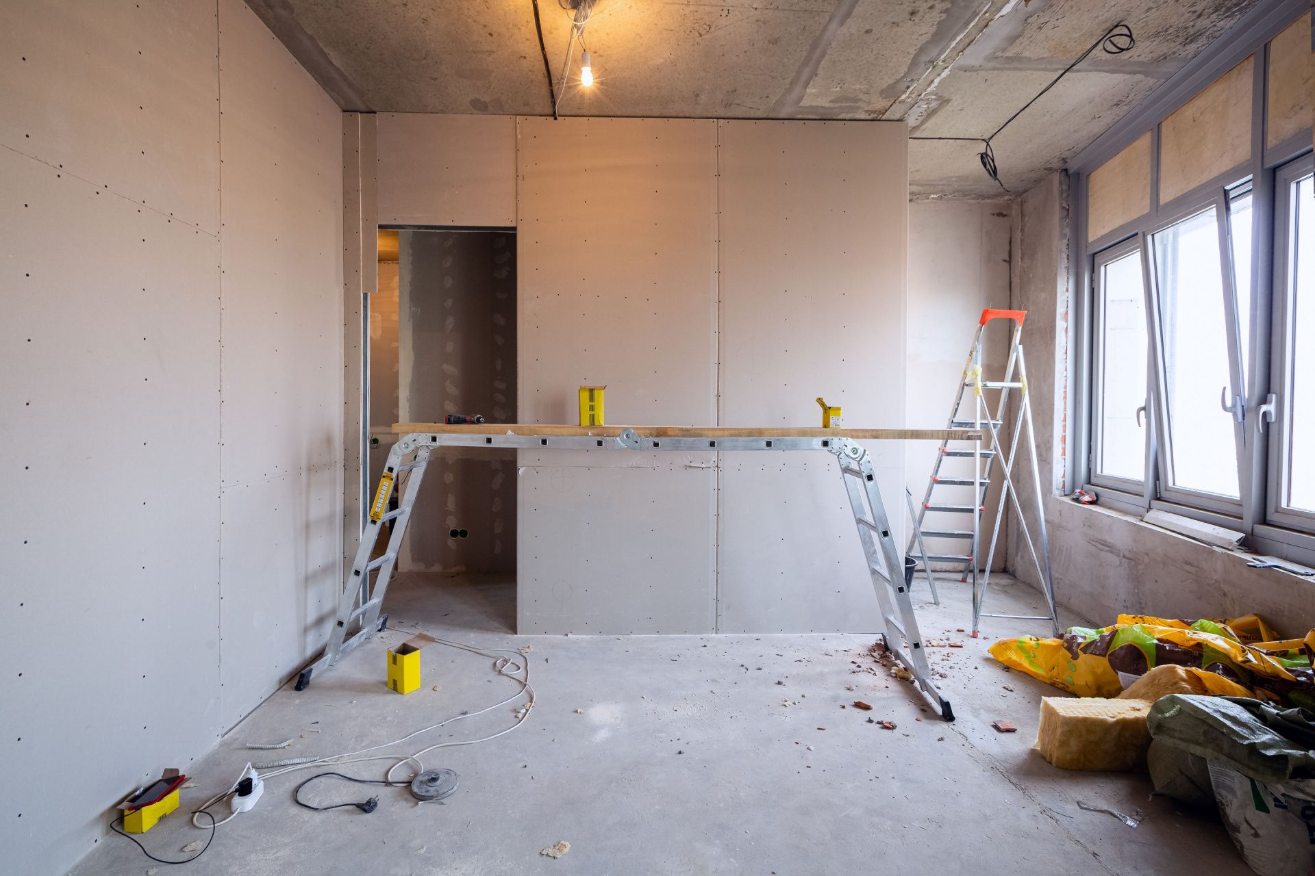 Room under construction, drywall, ladders, and tools present. Concrete floor, unfinished windows.