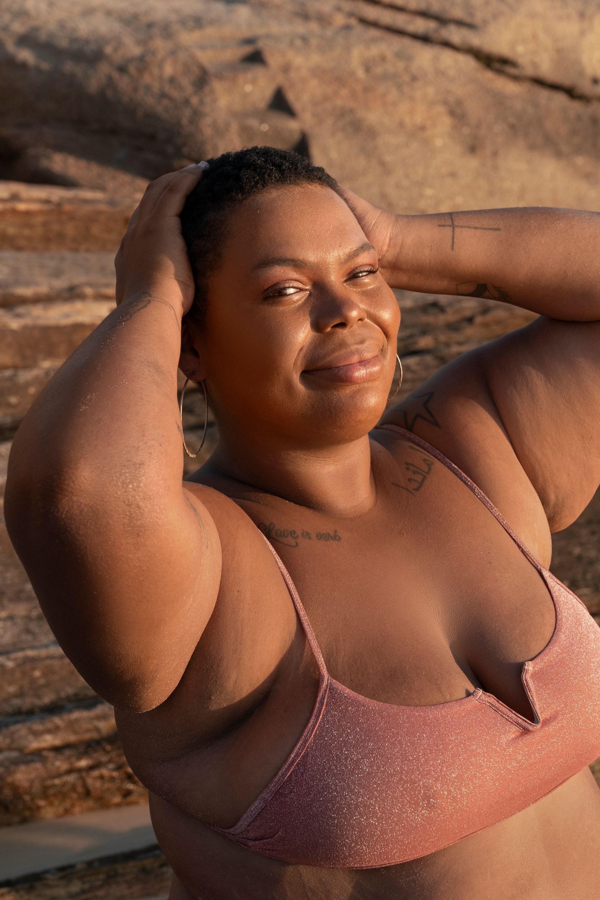 A woman in a bikini top is standing on a beach with her hands on her head.