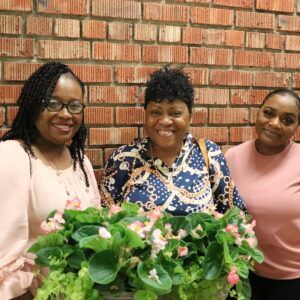 Three women are posing for a picture with a potted plant in front of a brick wall.