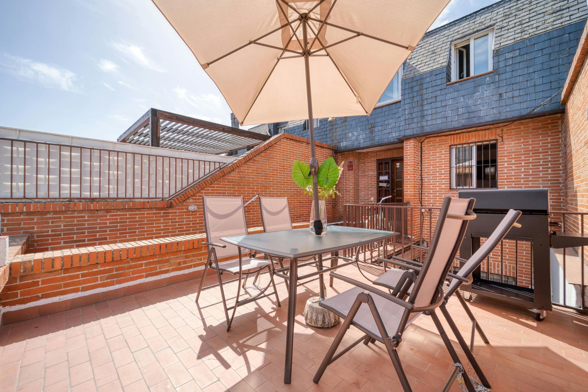 A brick terrace featuring a metal table, four chairs, a beige umbrella, and a barbecue grill under a clear blue sky.