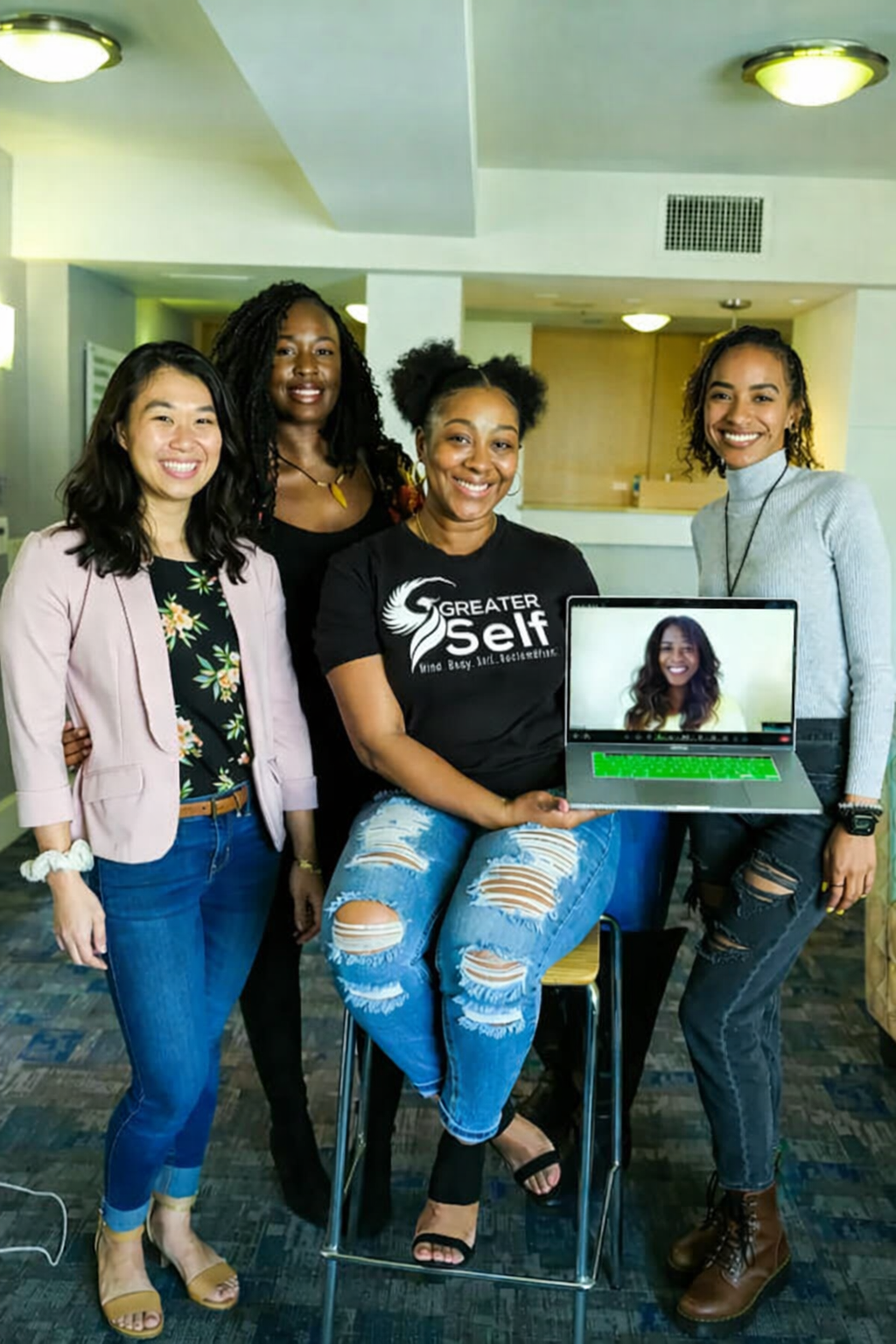 Four people, one holding a laptop, smiling at the camera. They are in a room with a light and window.
