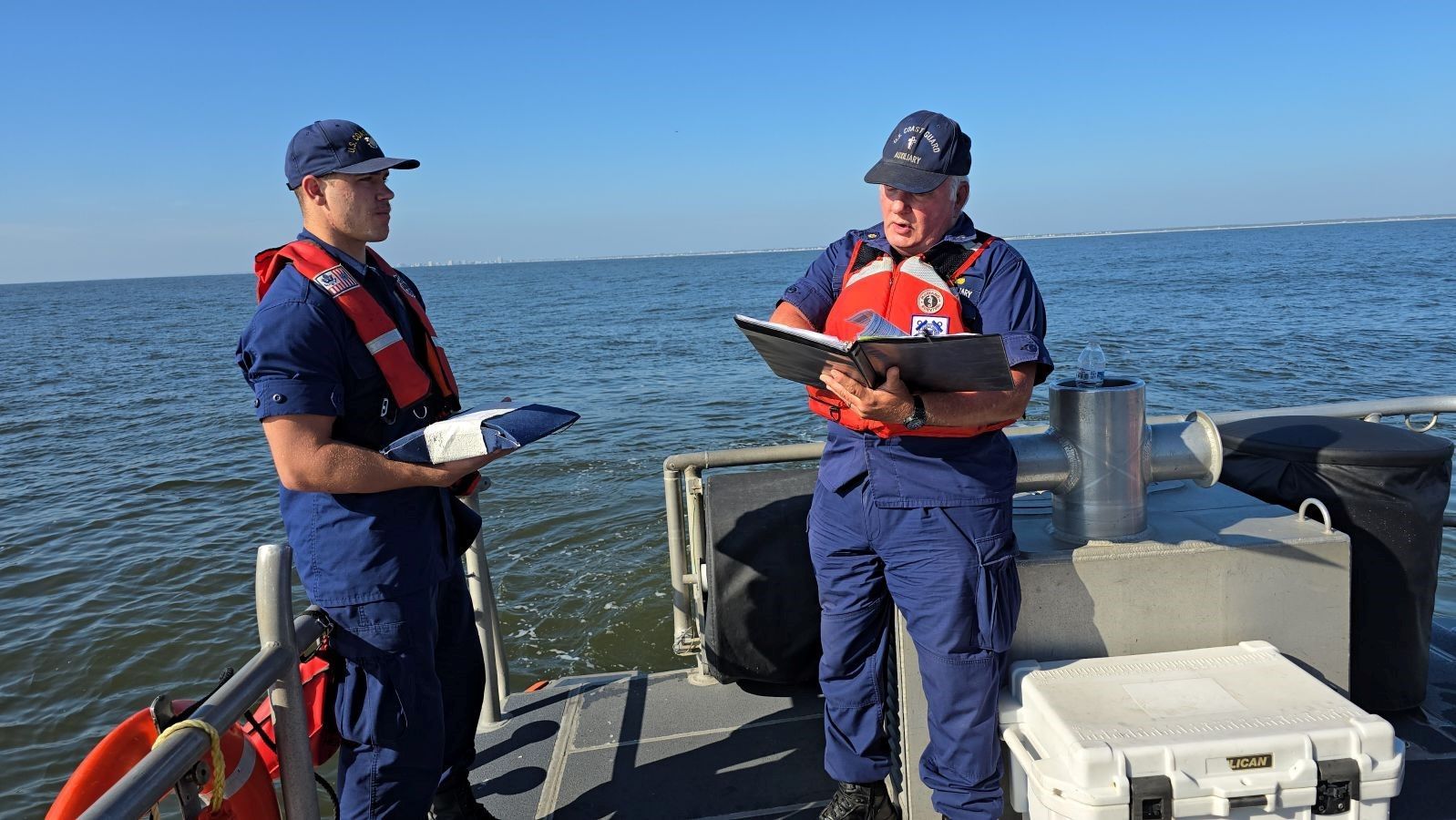 Two coast guard officers are standing on a boat looking at a clipboard.