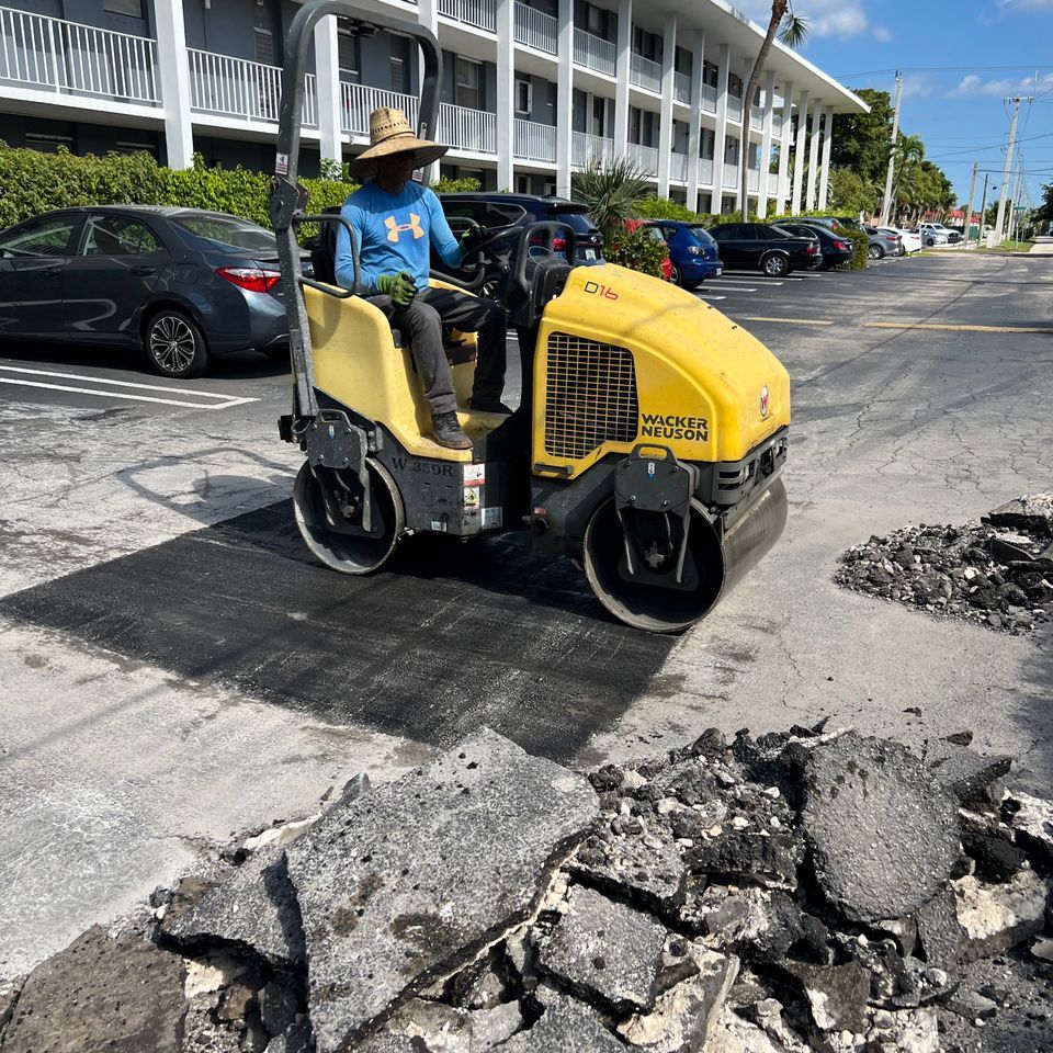 Person operating a yellow asphalt roller on a freshly paved road section, next to a pile of old asphalt.