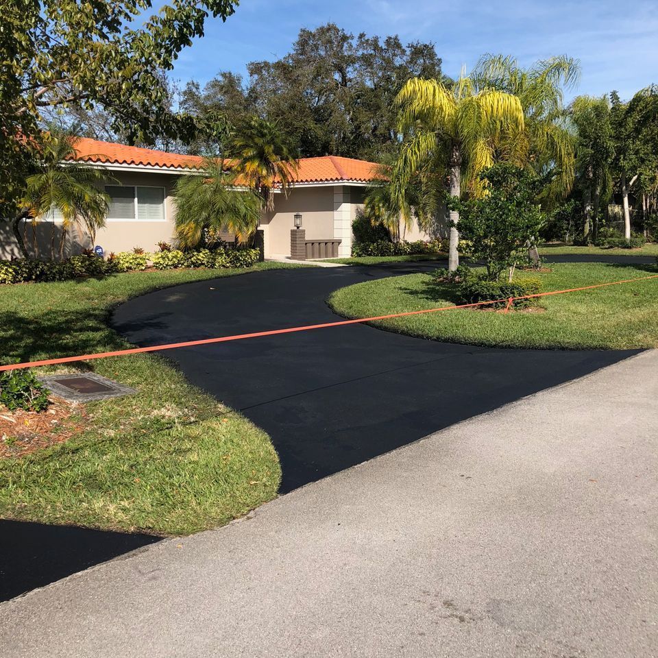 Newly sealed black asphalt driveway curving to a beige house with an orange tile roof, surrounded by green grass and trees.