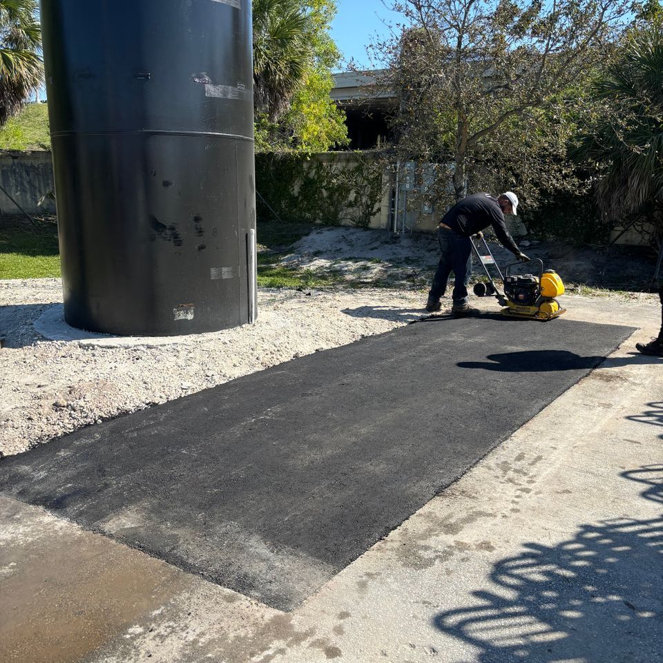 Worker compacting fresh asphalt path beside a black tank. White gravel border. Outdoors.