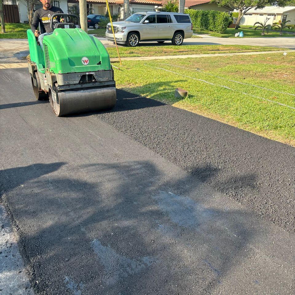 Man operating green asphalt roller compacting freshly laid black asphalt on a residential street.